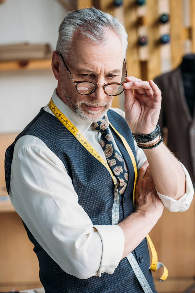 handsome tailor touching glasses and looking down at sewing workshop - Photo, Image