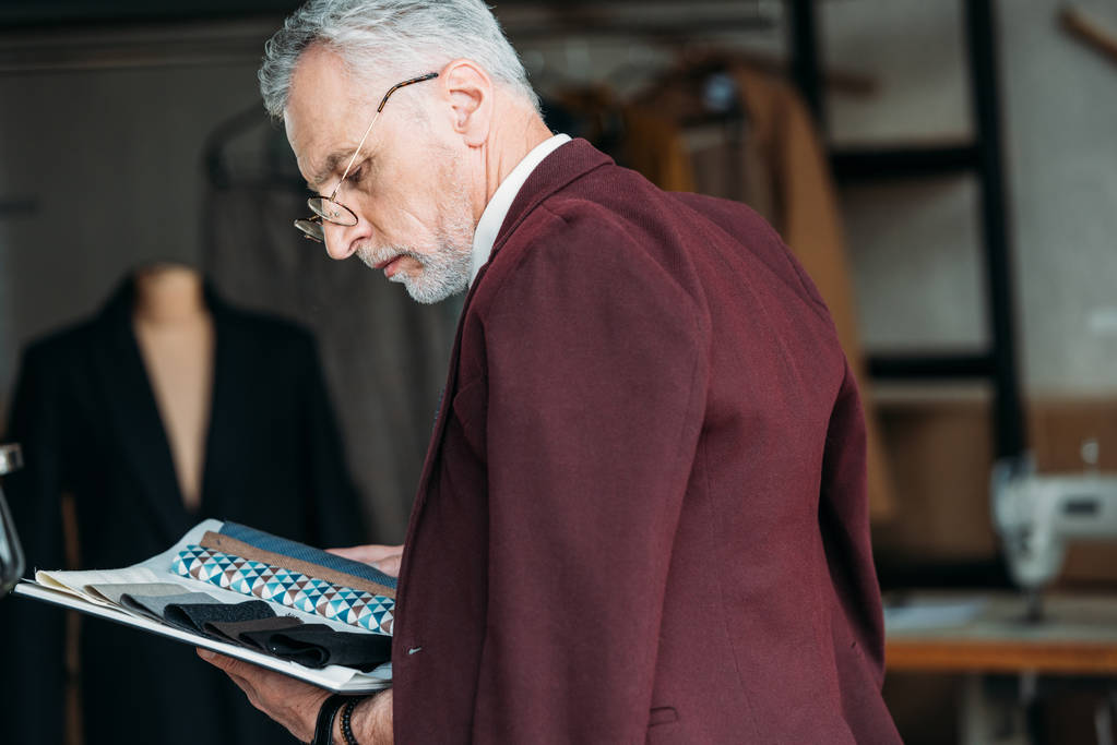 alfaiate maduro concentrado com catálogo de tipos de pano e jaqueta nos ombros na oficina de costura
 - Foto, Imagem