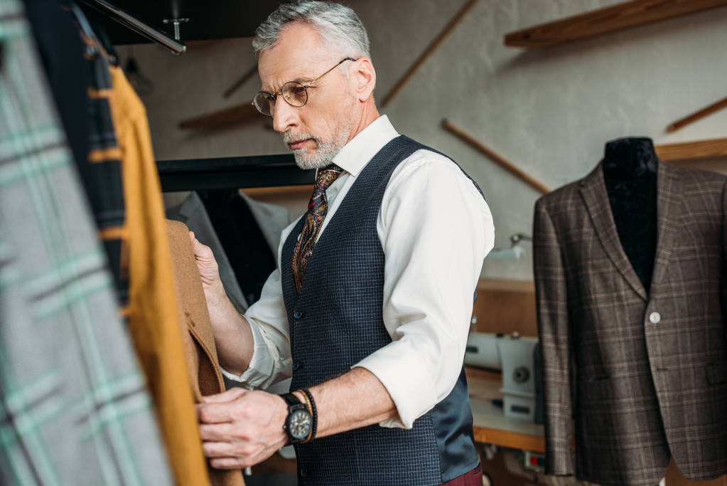 handsome mature tailor examining jackets at sewing workshop - Photo, Image
