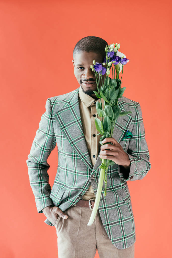 улыбающийся африканский американец в куртке, держащий букет цветов, изолированный на красном
 - Фото, изображение
