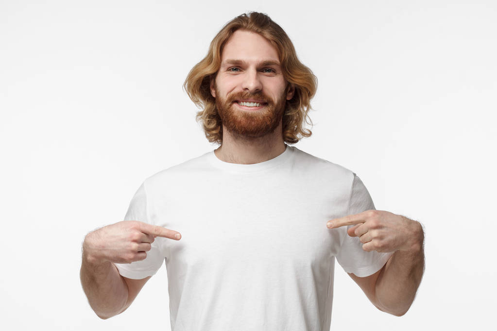 Горизонтальный крупный план красивого бородатого парня со светлыми волосами и усами на белом фоне, указывающий на белую пустую футболку, которую он носит обеими руками, улыбающийся, копирайт для рекламы
 - Фото, изображение