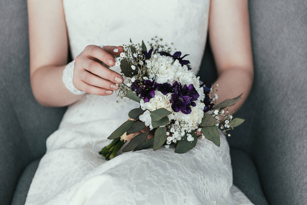 частичный вид невесты в белом платье с красивым свадебным букетом, отдыхающей в кресле
 - Фото, изображение