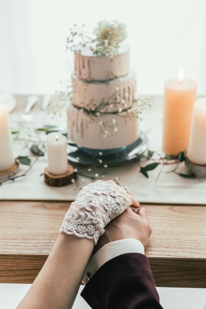 gedeeltelijke weergave van de pasgetrouwden hand in hand met bruidstaart, rustieke bruiloft concept zittend aan tafel geserveerd - Foto, afbeelding