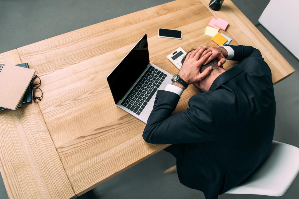 hoge hoekmening van gestresste zakenman met handen op kop liggen op de werkplek met laptop in kantoor - Foto, afbeelding