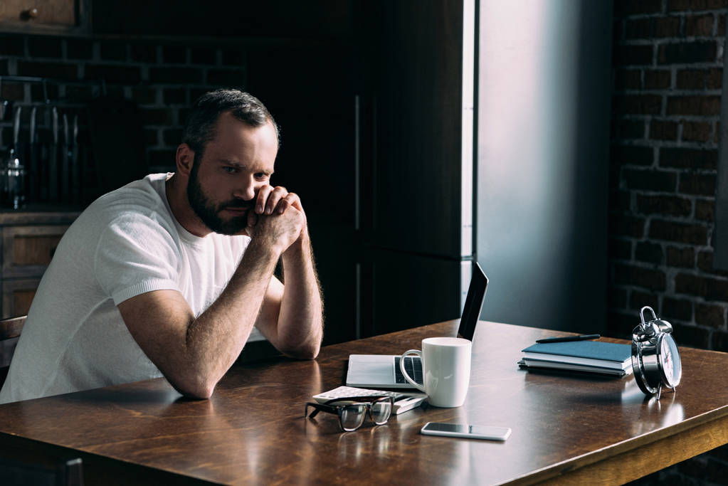 κατάθλιψη νεαρός άνδρας κάθεται στην κουζίνα με το laptop και αναζητούν μακριά - Φωτογραφία, εικόνα