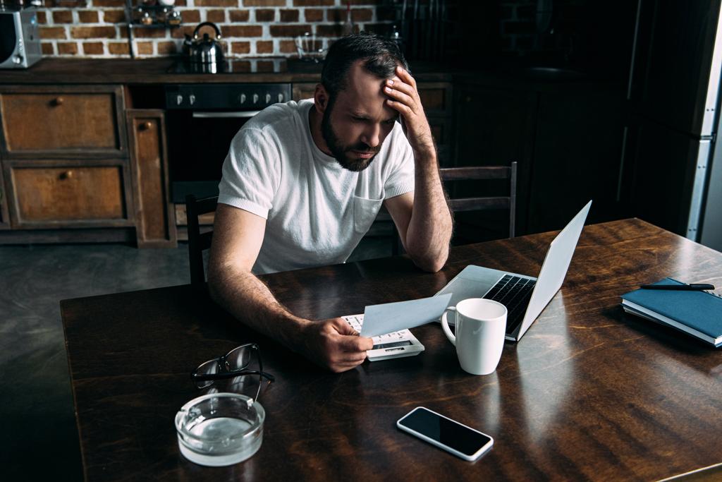 depressziós fiatal férfi nézett fotó-kártya a kezében laptop konyha ülve - Fotó, kép