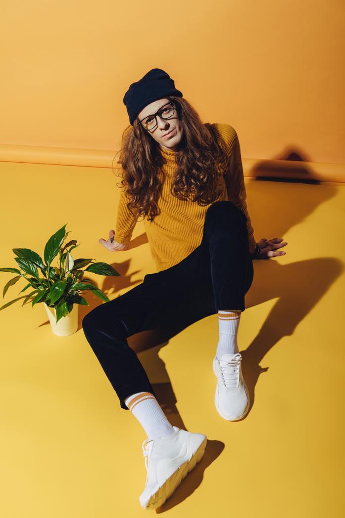 modieuze man met lange haren zitten in de buurt van kamerplant, op geel - Foto, afbeelding