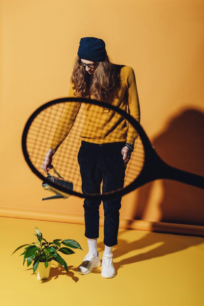 stylish man watering plant, view through tennis racket on yellow - Photo, Image
