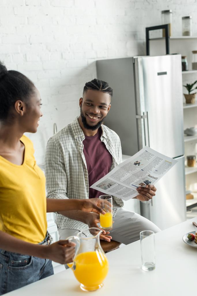 アフリカ系アメリカ人彼氏読書新聞とキッチンでガラスにオレンジ ジュースを注ぐのガール フレンドを笑顔 - 写真・画像