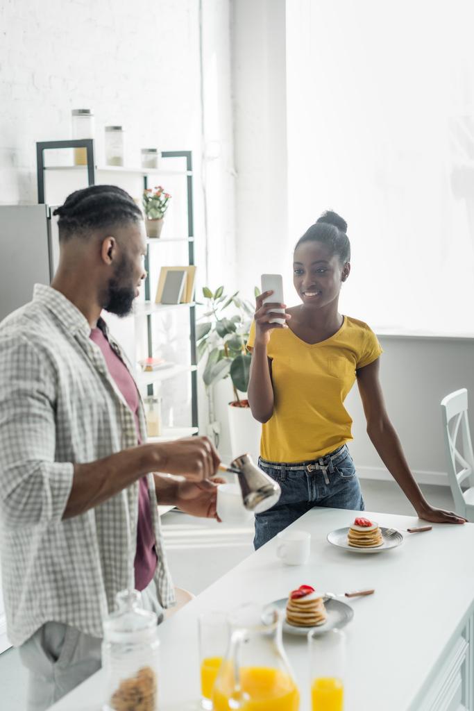 afro-americana namorada tirar foto de namorado com café com smartphone na cozinha
 - Foto, Imagem