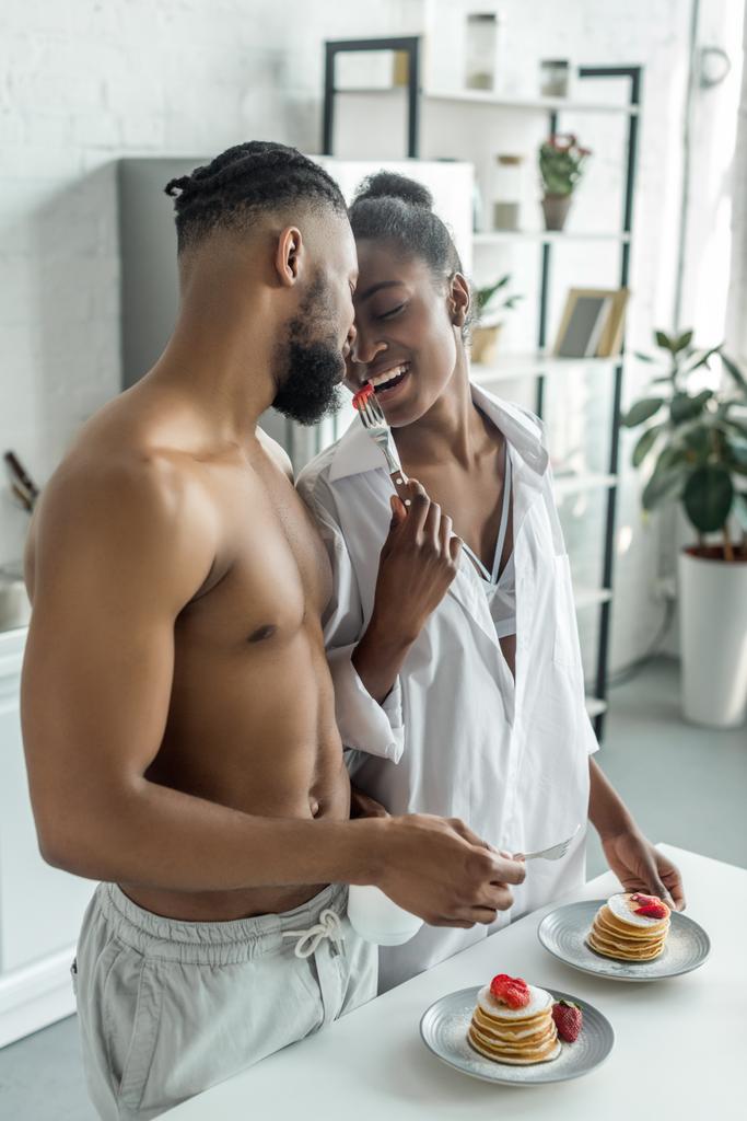 african american girlfriend holding fork with strawberry between her and boyfriend at kitchen - Photo, Image