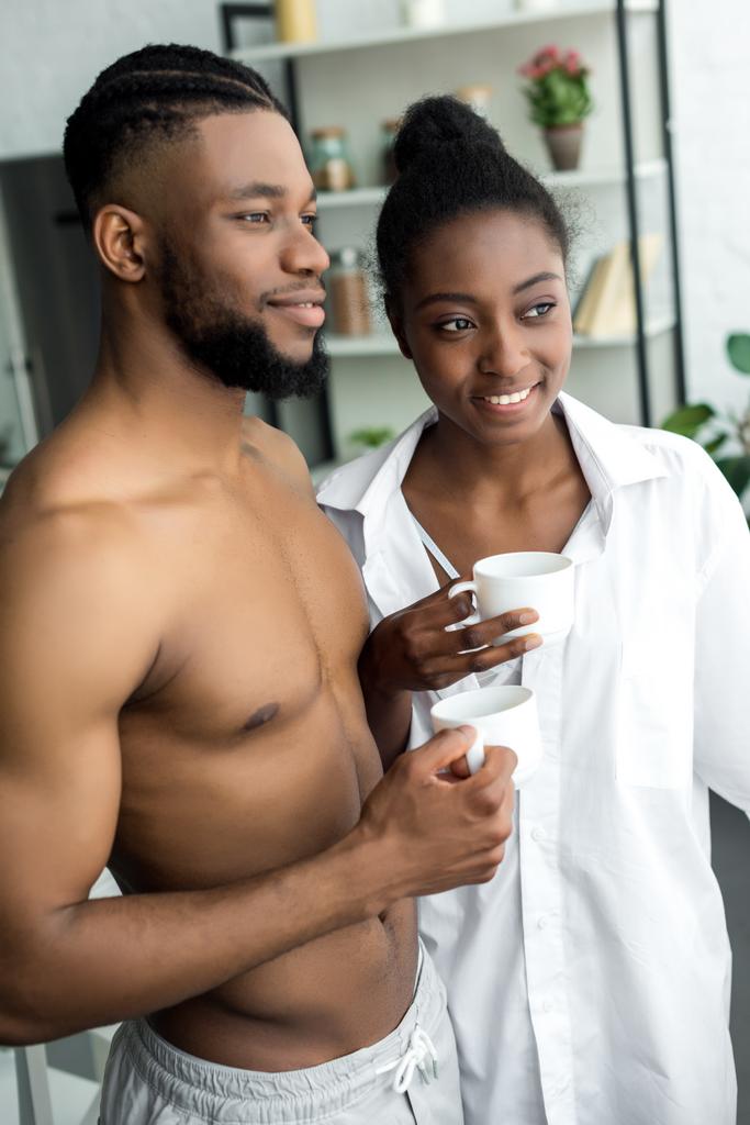african american couple holding cups and looking away at kitchen - Photo, Image