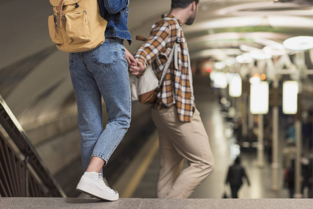 tiro recortado de casal de turistas elegantes com mochilas de mãos dadas na estação de metrô
 - Foto, Imagem