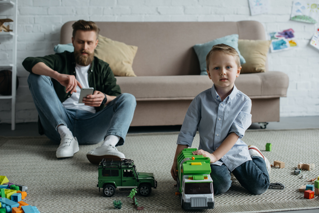 foco seletivo do pai com smartphone e filho pequeno com carros de brinquedo no chão em casa
 - Foto, Imagem