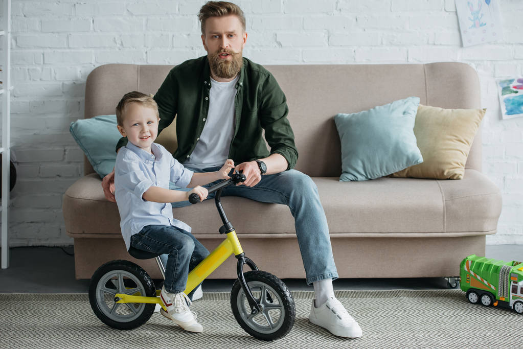 klein kind op evenwicht fiets met bebaarde vader zittend op de Bank thuis - Foto, afbeelding
