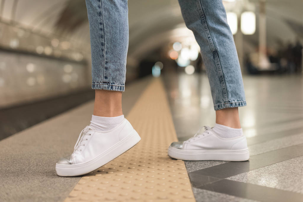 περικομμένη εικόνα γυναικεία πόδια σε κομψά τζιν και αθλητικά παπούτσια στο σταθμό του μετρό  - Φωτογραφία, εικόνα