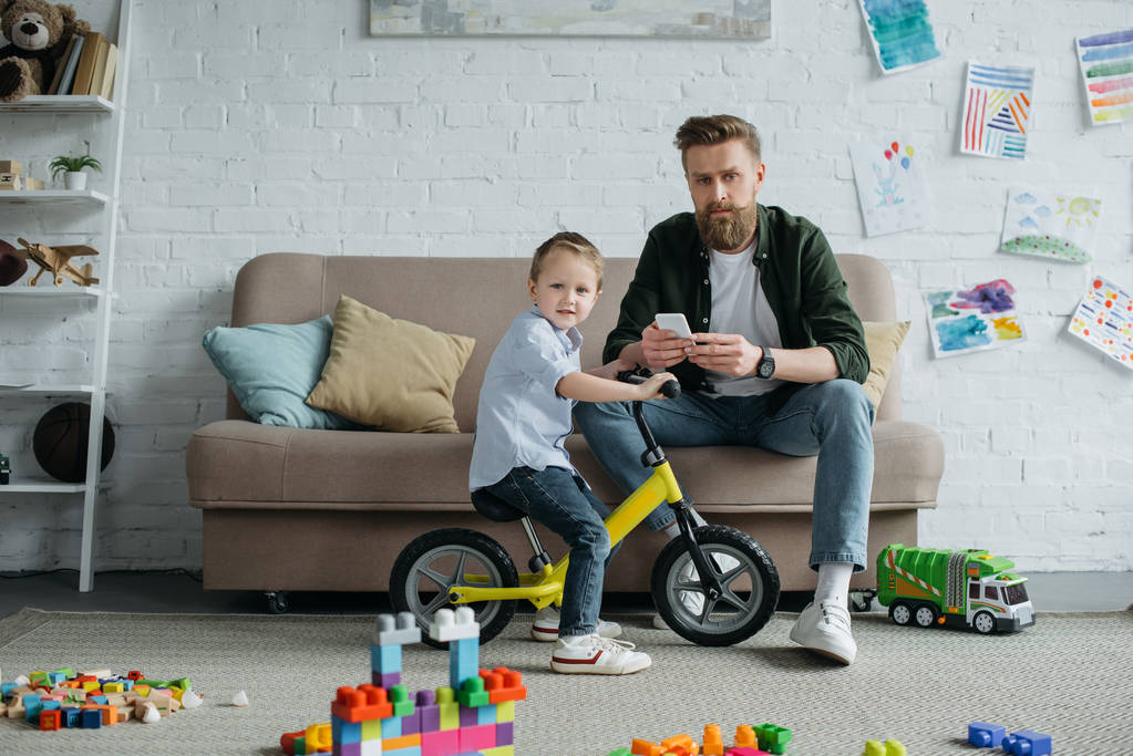 маленький ребенок на балансе велосипед с бородатым отцом со смартфоном сидит на диване дома
 - Фото, изображение