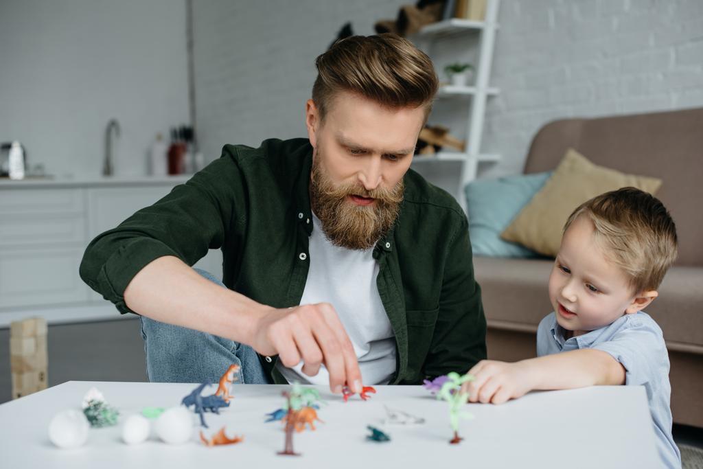 Vater und süßer kleiner Sohn spielen zu Hause mit verschiedenen Spielzeug-Dinosauriern - Foto, Bild
