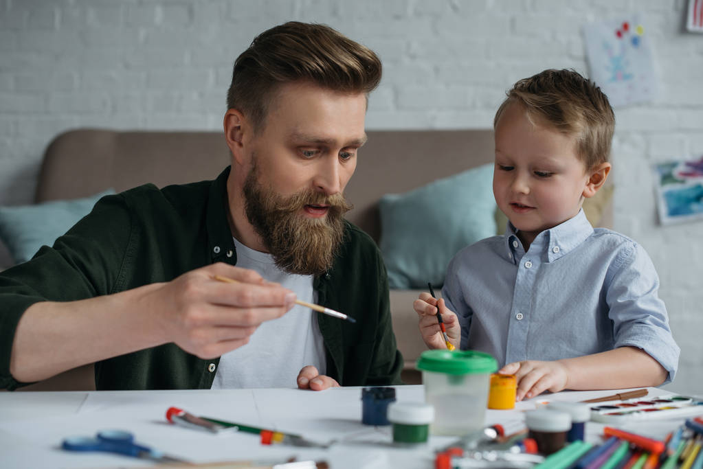 портрет отца и милого маленького сына с красками и кисточками, рисующих картины вместе дома
 - Фото, изображение