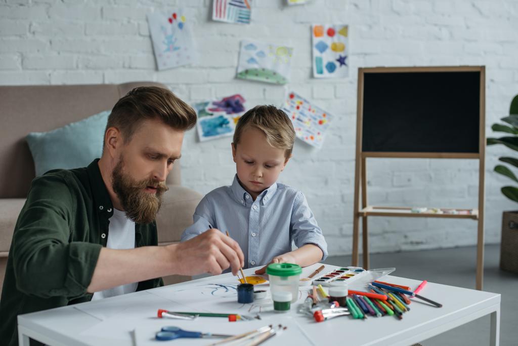 focused father and cute little son with paints and brushes drawing pictures together at home - Photo, Image