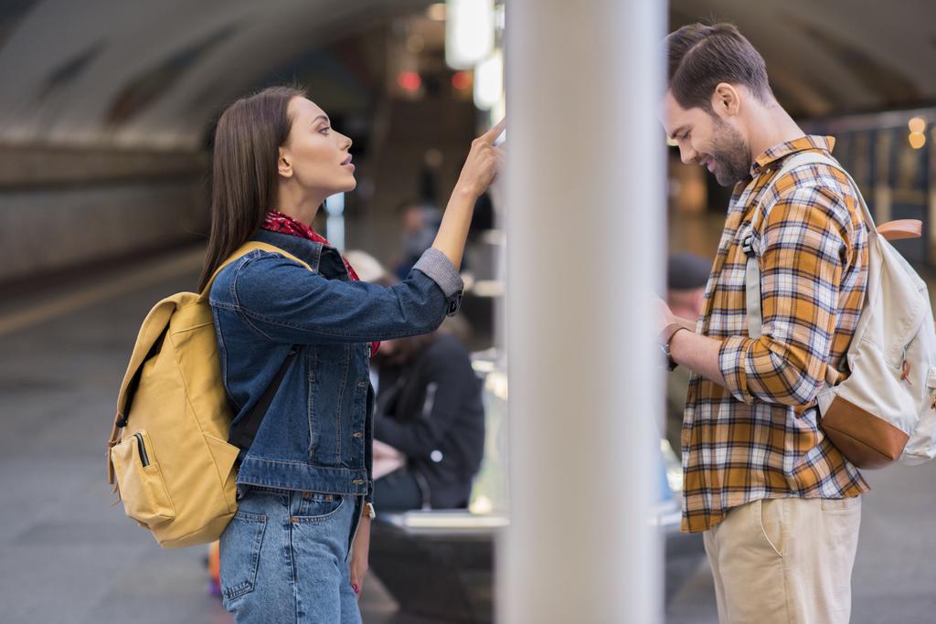 vue latérale du voyageur féminin regardant le panneau d'information et le petit ami en utilisant un smartphone à la station de métro
  - Photo, image