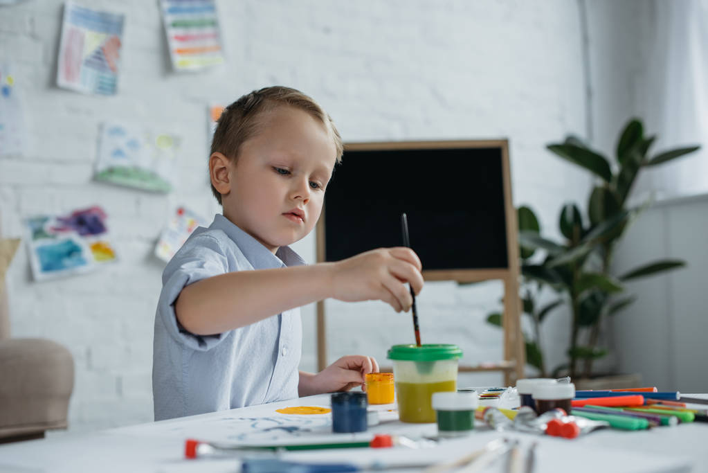 ориентированный маленький мальчик с кистью и картинами рисует картину в одиночестве дома
 - Фото, изображение