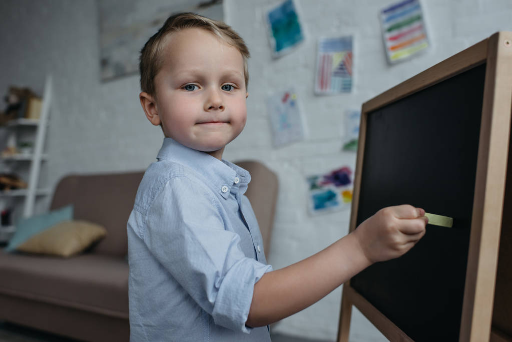 vista lateral del niño pequeño con un pedazo de tiza mirando a la cámara mientras dibuja la imagen en la pizarra en casa
 - Foto, imagen