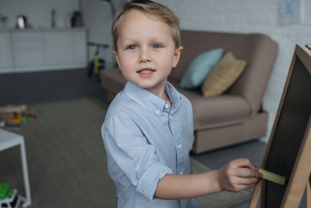 vista lateral del niño pequeño con un pedazo de tiza mirando a la cámara mientras dibuja la imagen en la pizarra en casa
 - Foto, Imagen