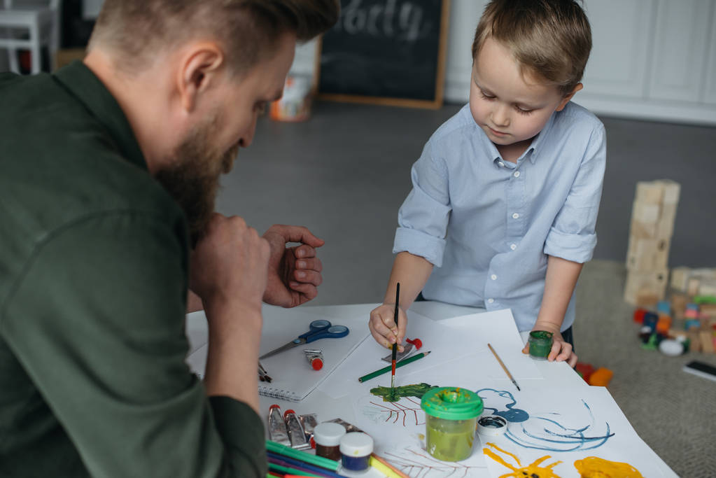 маленький мальчик с кистью и красками рисует картину вместе с отцом дома
 - Фото, изображение