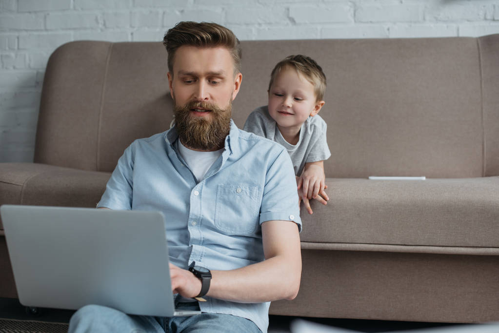 parrakas mies, joka käyttää kannettavaa tietokonetta poikansa kanssa lähellä sohvalla kotona
 - Valokuva, kuva