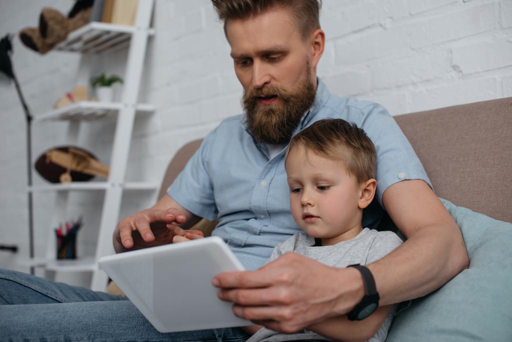 сфокусированные отец и маленький сын, используя планшет вместе на диване дома
 - Фото, изображение