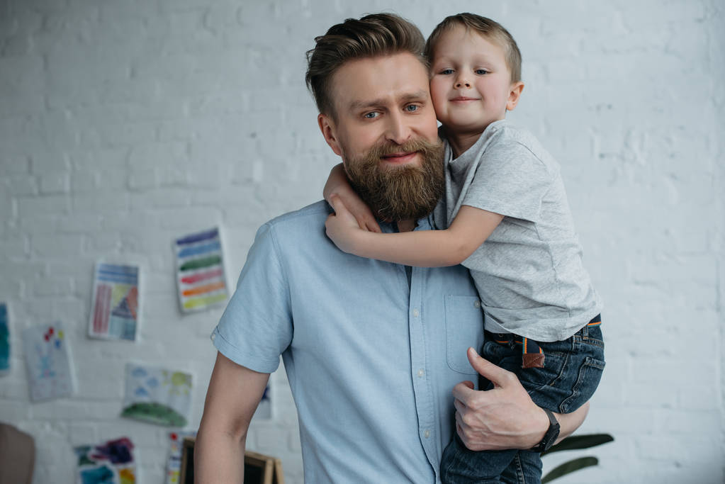 portrait of smiling father holding little son and looking at camera at home - Photo, Image