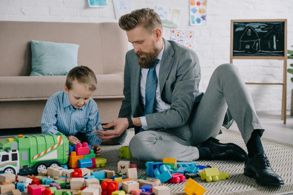 Geschäftsmann im Anzug und kleiner Sohn spielen zu Hause mit bunten Klötzen auf dem Boden, Arbeits- und Life-Balance-Konzept - Foto, Bild