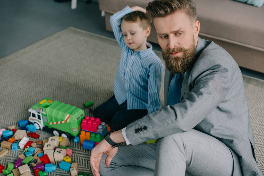 επιλεκτική εστίαση συλλογισμένος επιχειρηματία στο κοστούμι κάθεται κοντά ο μικρός γιος στο πάτωμα στο σπίτι, εργασία και τη ζωή ισορροπία έννοια - Φωτογραφία, εικόνα