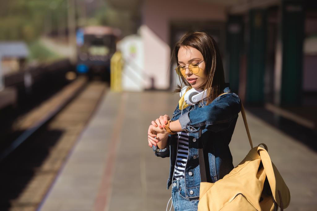κομψή γυναίκα σε γυαλιά ηλίου με ακουστικά και σακίδιο βλέπουν Ρολόι χεριού  - Φωτογραφία, εικόνα