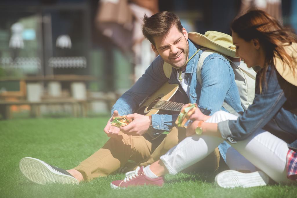 sonriente pareja de viajeros con la guitarra comiendo sándwiches y sentado en la hierba
  - Foto, imagen