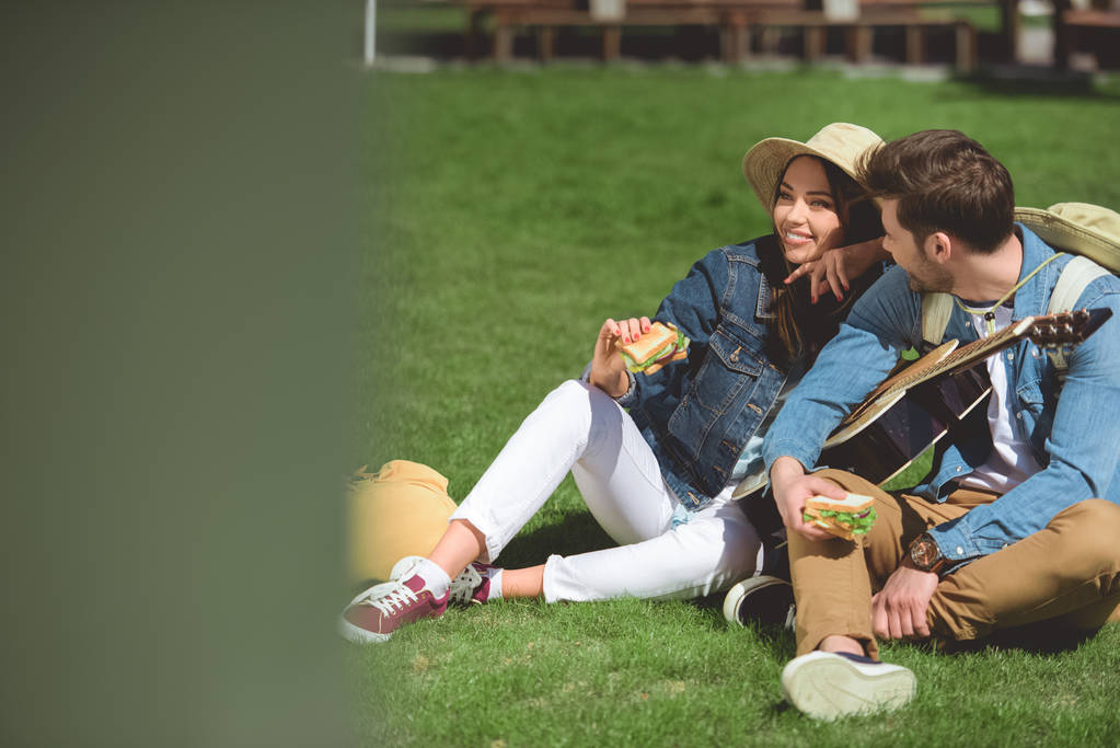 smiling couple of tourists in hats with guitar and sandwiches sitting on grass - Photo, Image