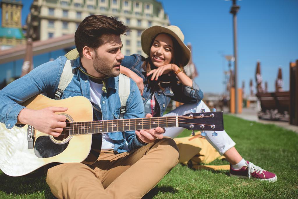 turista masculino tocando en la guitarra y novia mirándolo en la hierba
 - Foto, Imagen
