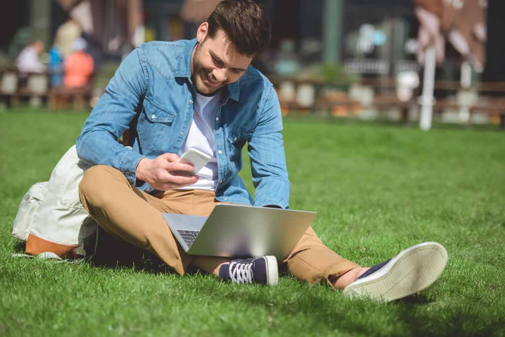 homem com laptop olhando para a tela do smartphone e sentado na grama
 - Foto, Imagem