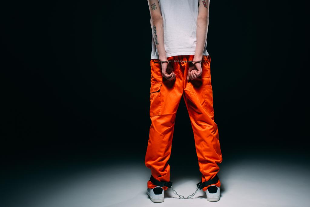 Vista recortada del hombre con uniforme de prisión con las manos esposadas detrás de la espalda sobre fondo oscuro
 - Foto, imagen
