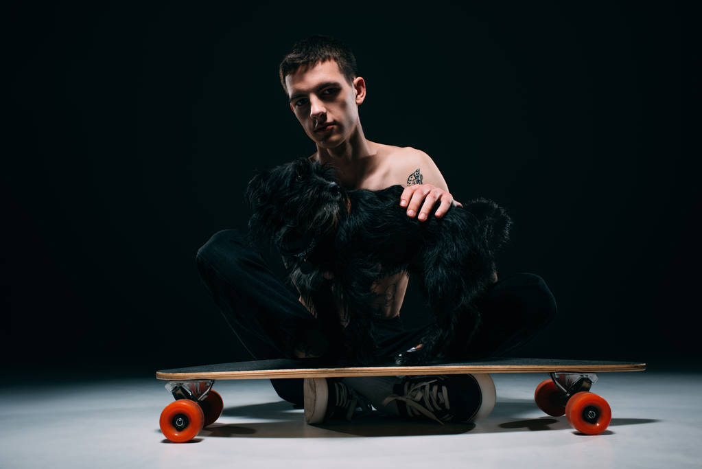 Man with tattoos stroking black dog on longboard on dark background - Photo, Image