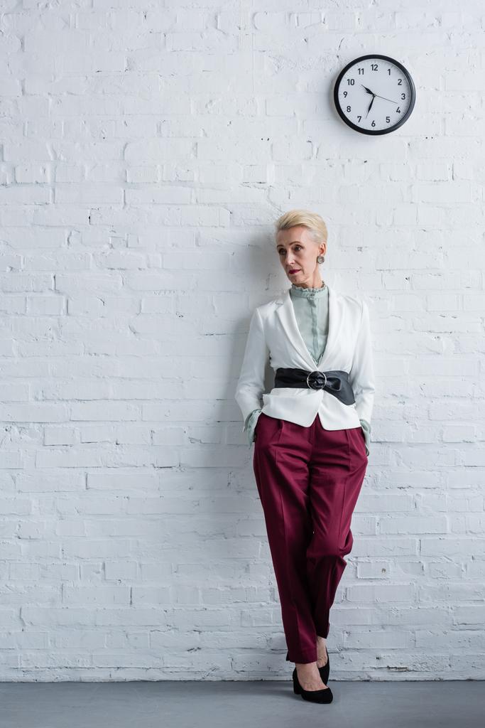 Elegancka bizneswoman starszy pozowanie na białej ścianie z zegarem - Zdjęcie, obraz