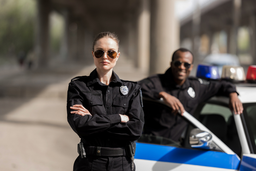 Młoda policjantka poważne, stojąc z skrzyżowanymi rękami podczas jej partner, stojący w pobliżu samochodu i uśmiechając się niewyraźne na tle - Zdjęcie, obraz