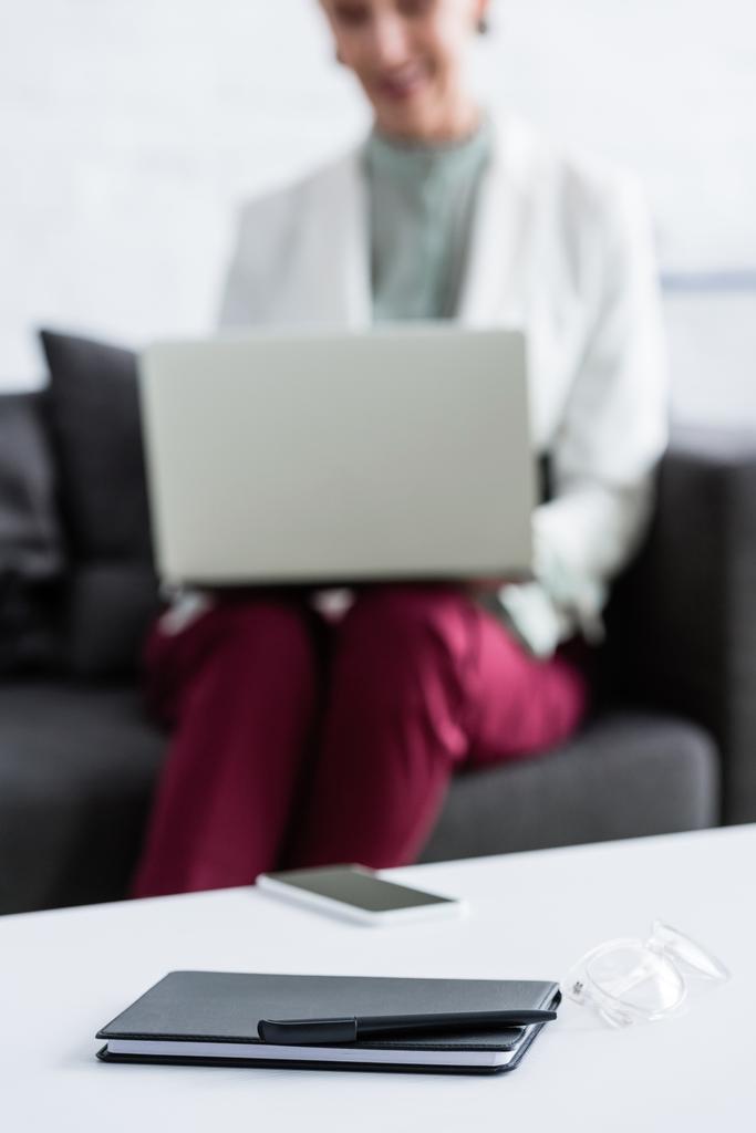 foco seletivo da mulher de negócios usando laptop, smartphone, diário, caneta e óculos deitados na mesa em primeiro plano
 - Foto, Imagem