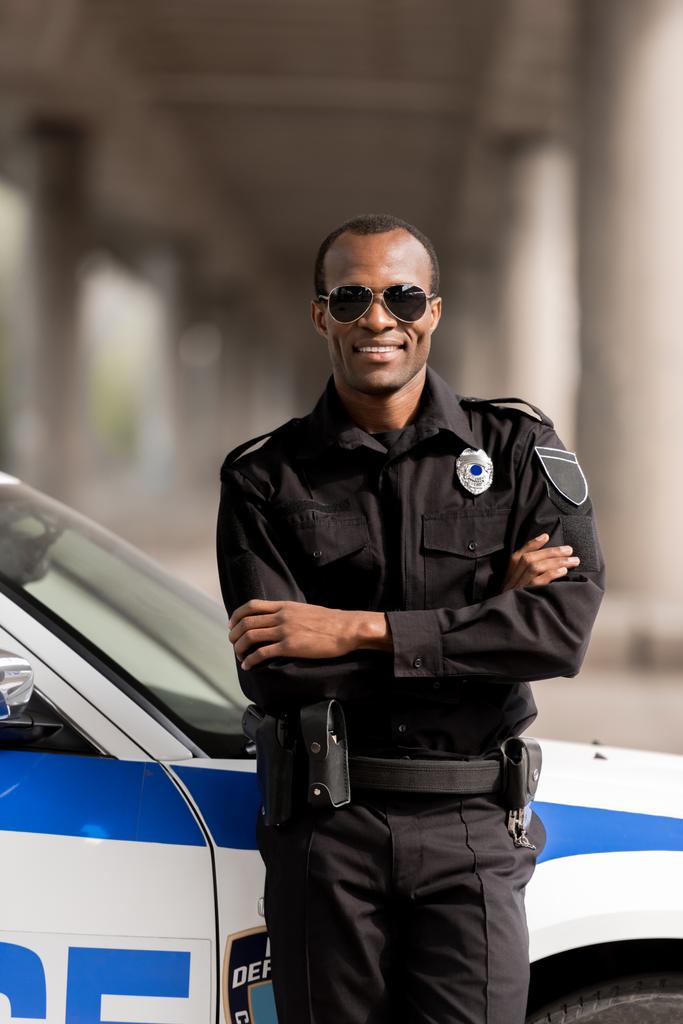 lächelnder afrikanisch-amerikanischer Polizist mit verschränkten Armen, der sich auf sein Auto zurücklehnt und in die Kamera blickt - Foto, Bild