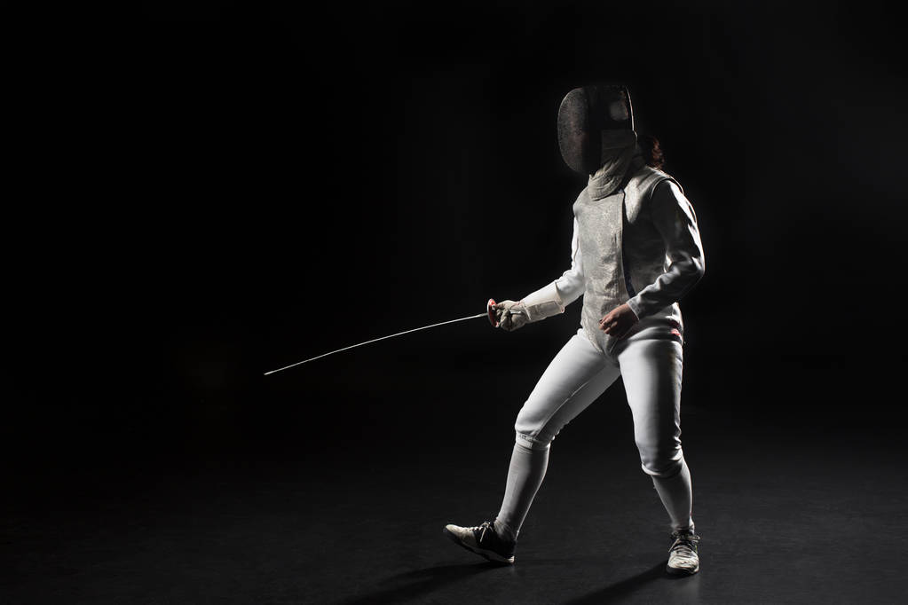 full length of female fencer in uniform posing with sword on black background - Photo, Image