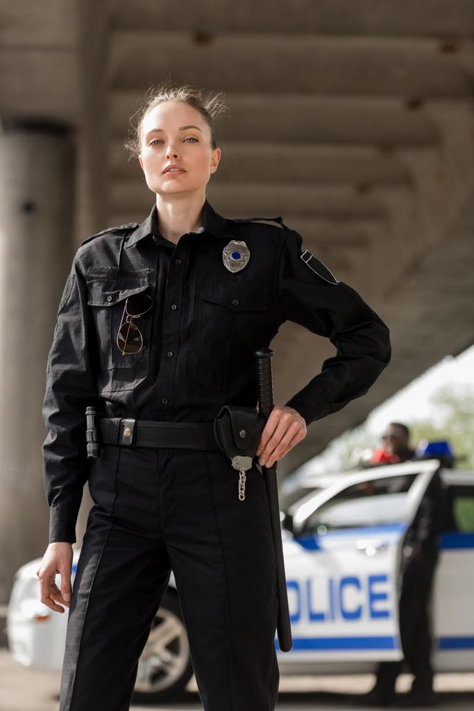 atractiva oficial de policía femenina en uniforme mirando a la cámara con compañero borroso cerca del coche en el fondo
 - Foto, Imagen