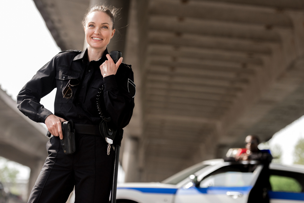 felice attraente poliziotta utilizzando walkie-talkie con partner offuscata vicino auto sullo sfondo
 - Foto, immagini