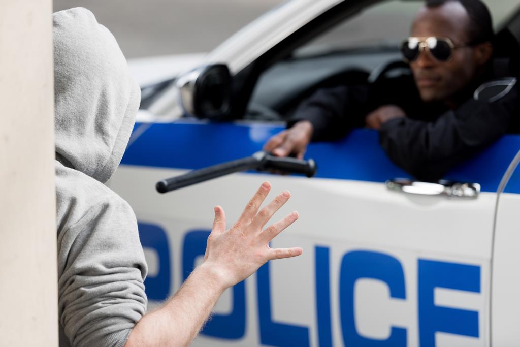 Polizist blickt aus Autoscheibe und zeigt mit Polizeiknüppel auf vermummten Mann - Foto, Bild