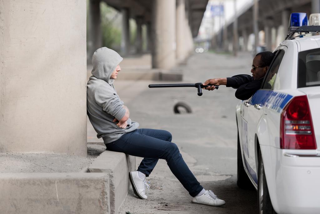 Polizist zeigt auf vermummten Mann mit Polizeiknüppel, während er auf der Straße sitzt - Foto, Bild